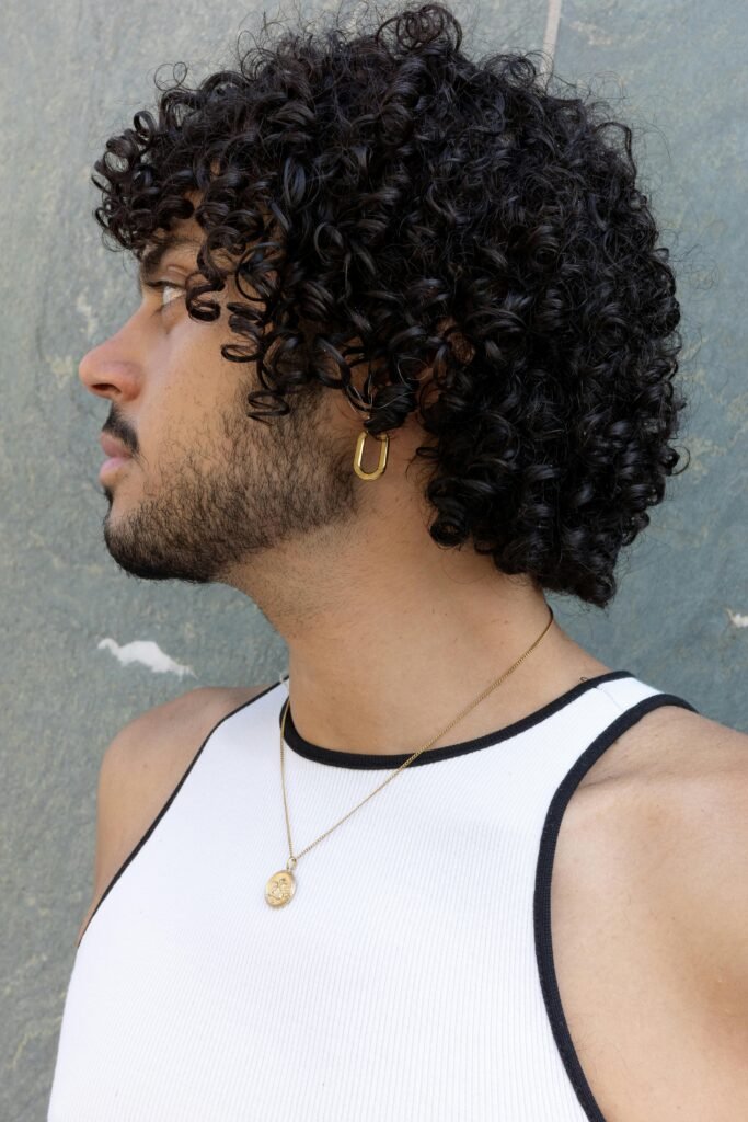 Side profile portrait of a young man with curly hair wearing a white tank top and gold jewelry.